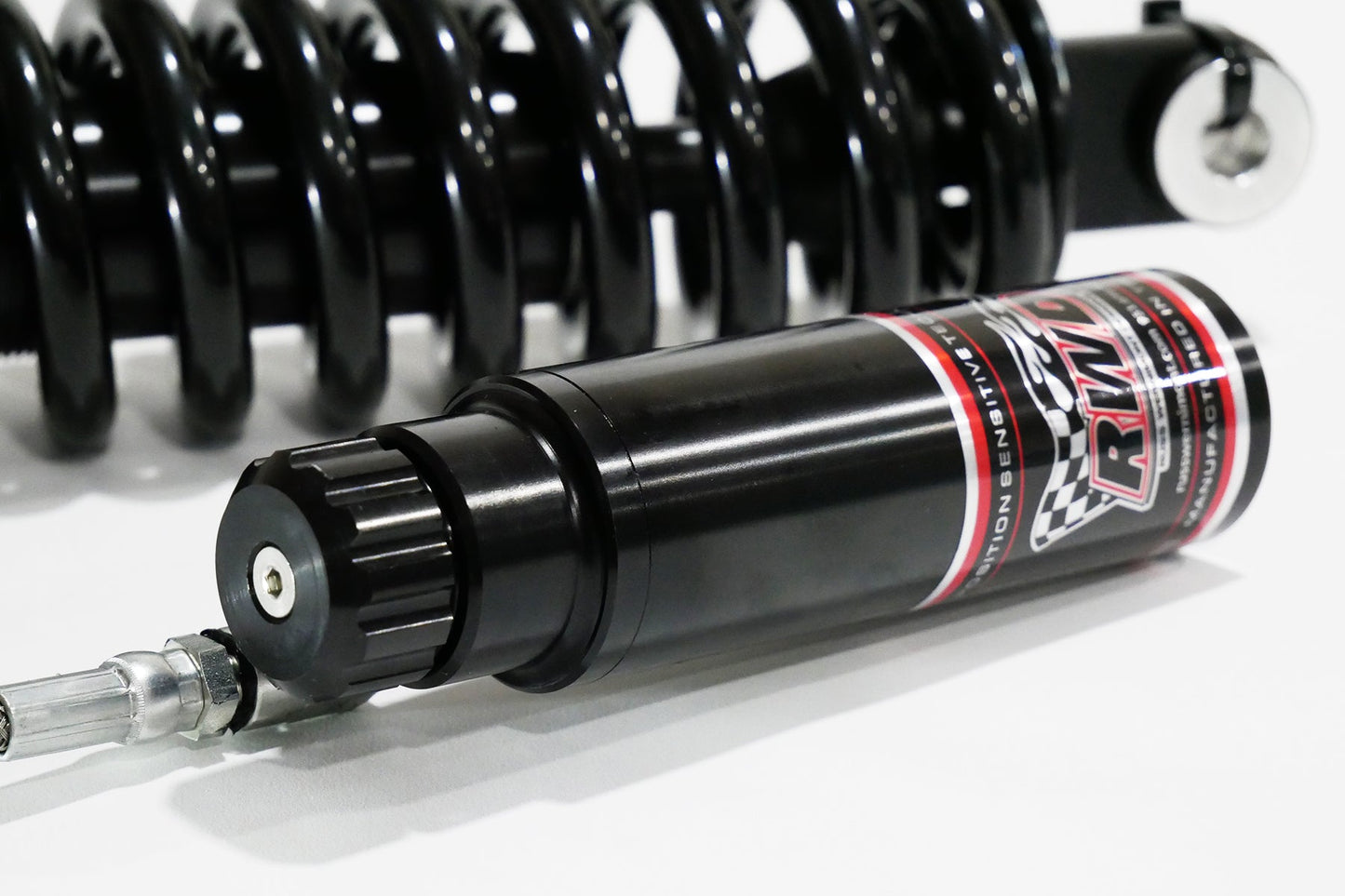 A close-up side shot of an RWD V-Twin RS-2 reservoir, featuring a black body with red and white RWD branding, connected to a silver hose, with a black spring in the background.